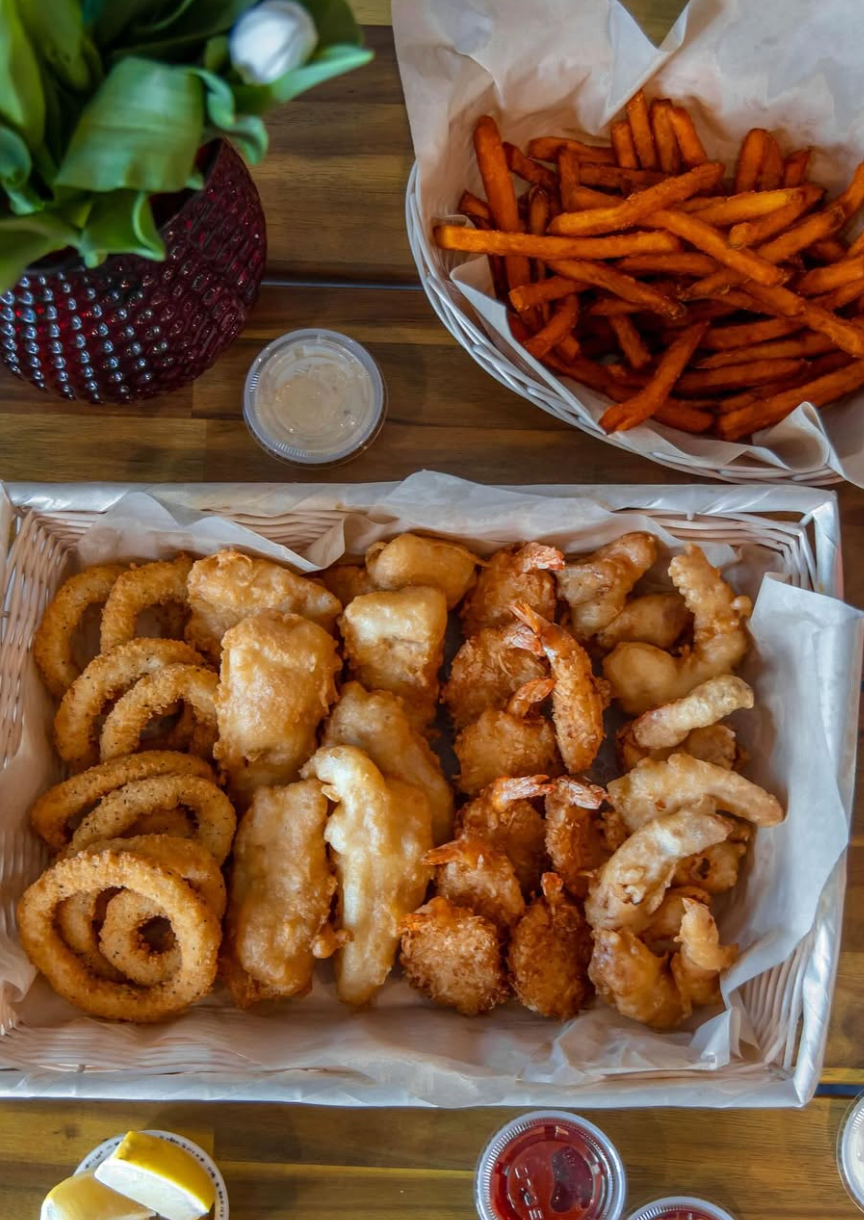 An overhead view of a seafood platter with golden-fried shrimp, fish, calamari, and onion rings in a lined basket, served alongside a basket of sweet potato fries, dipping sauces, and a small potted plant on a wooden table.