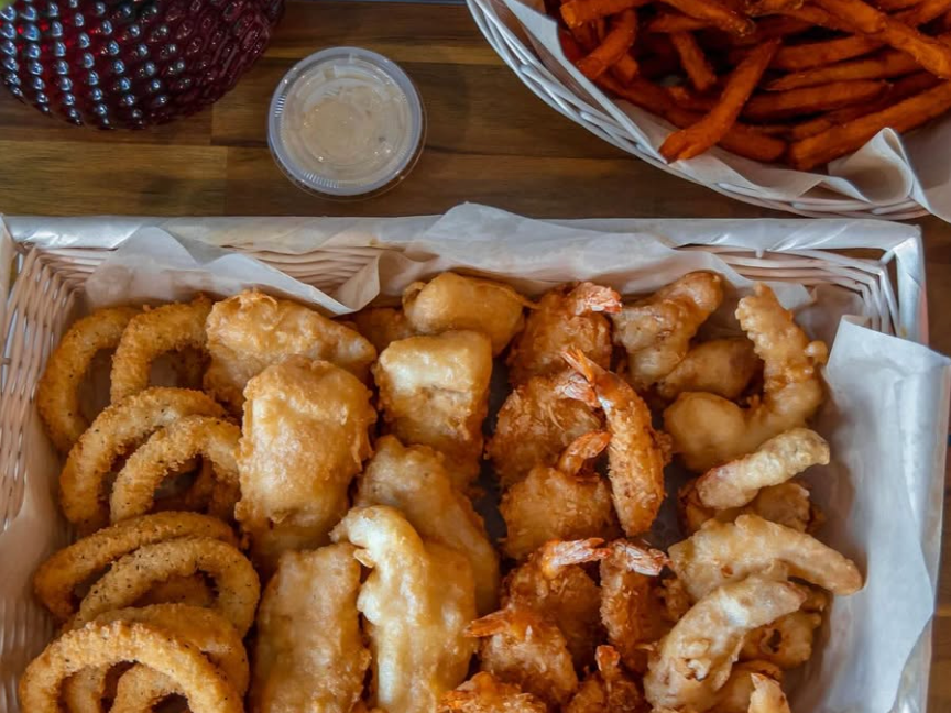 An overhead view of a seafood platter with golden-fried shrimp, fish, calamari, and onion rings in a lined basket, served alongside a basket of sweet potato fries, dipping sauces, and a small potted plant on a wooden table.
