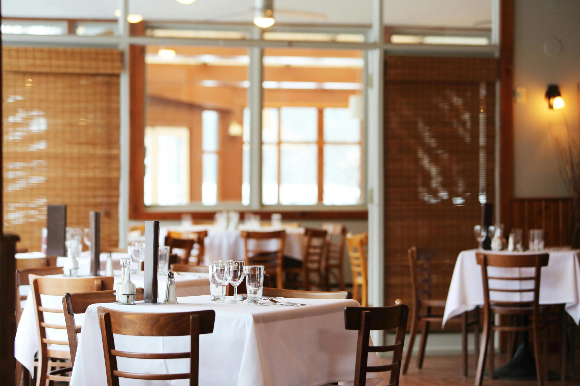 A warmly lit dining room with white tablecloths, wood chairs, and neatly set tables ready for guests. This peaceful restaurant space sets the tone for intimate meals and quiet conversation.