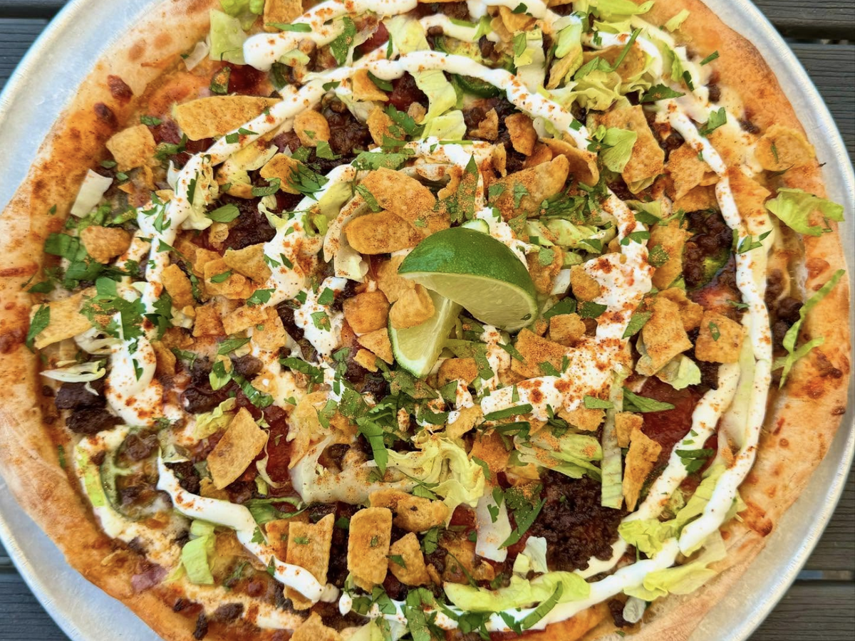 A taco-inspired pizza topped with seasoned ground beef, shredded lettuce, corn chips, sour cream drizzle, fresh cilantro, and lime wedges, served on a metal tray.