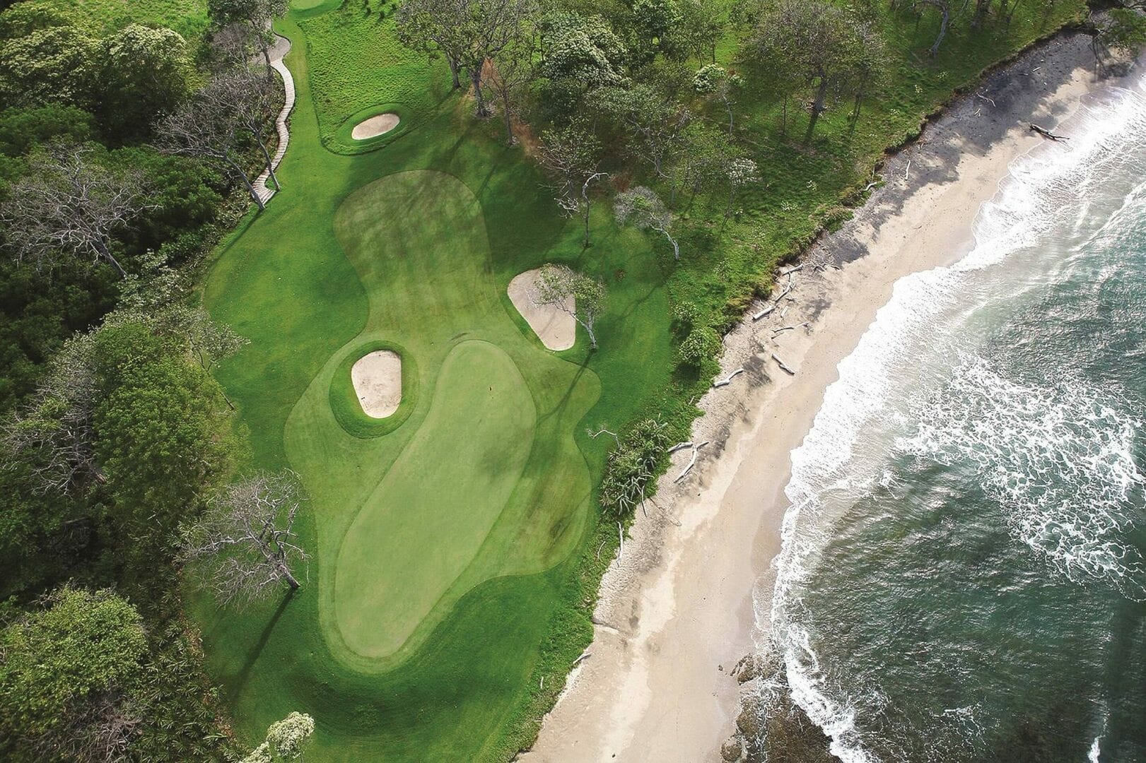 An aerial view of a lush golf course in Tamarindo, Costa Rica, set right beside a quiet sandy beach and rolling ocean waves. Bright green fairways and sand bunkers sit just steps from the shoreline, surrounded by tropical trees. Tamarindo is known for its surf spots, warm weather, and scenic coastal golf views.