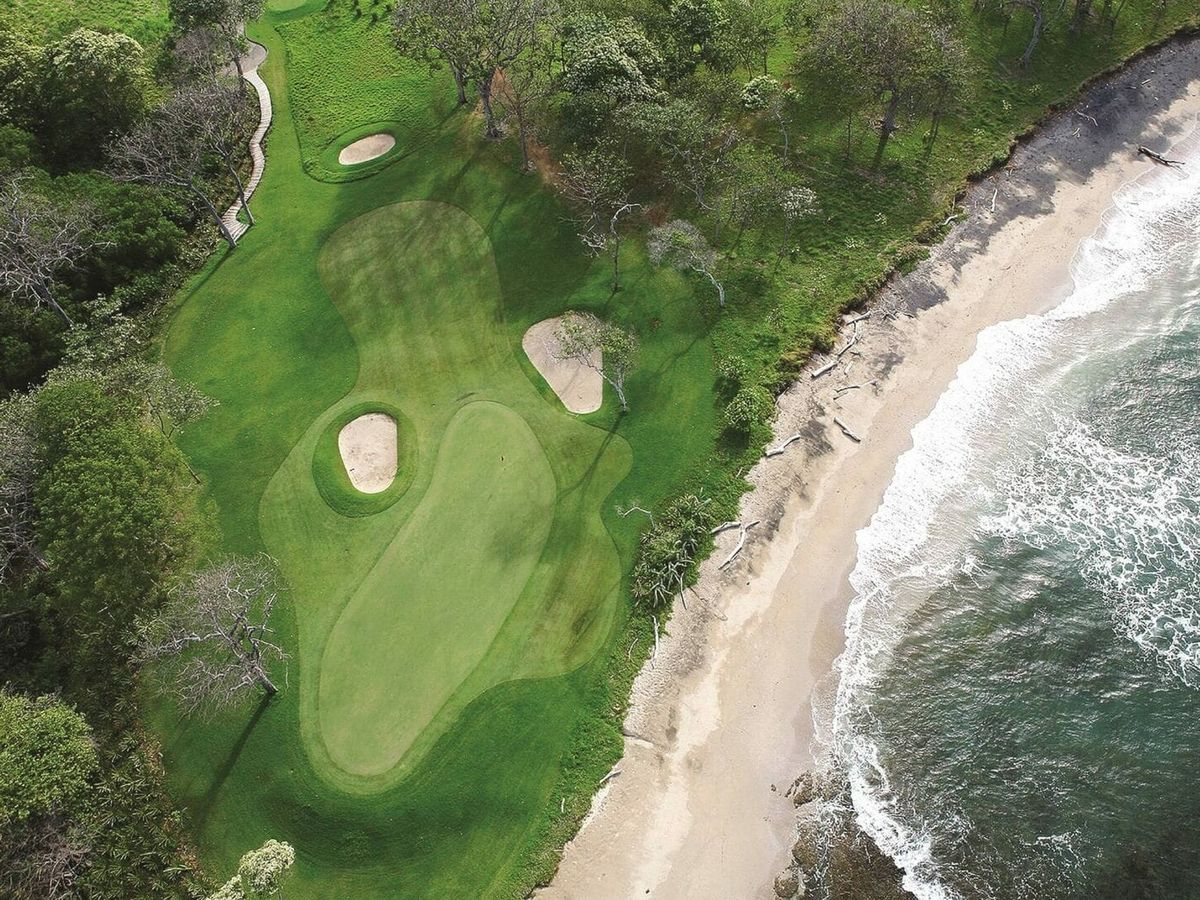 An aerial view of a lush golf course in Tamarindo, Costa Rica, set right beside a quiet sandy beach and rolling ocean waves. Bright green fairways and sand bunkers sit just steps from the shoreline, surrounded by tropical trees. Tamarindo is known for its surf spots, warm weather, and scenic coastal golf views.