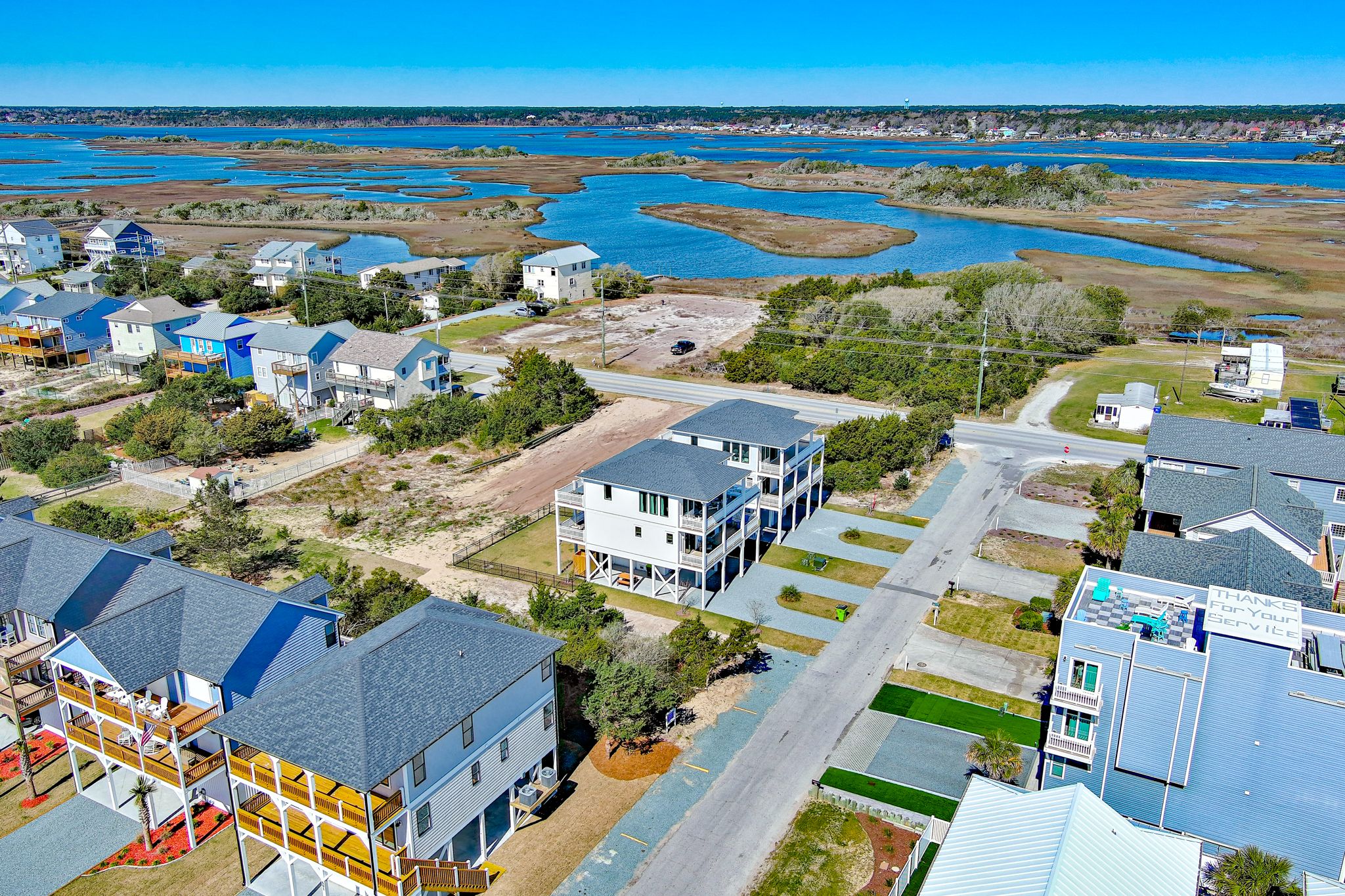 Aerial view of a coastal neighborhood featuring elevated beach homes, a winding marshland with blue water in the background, and a clear blue sky above.