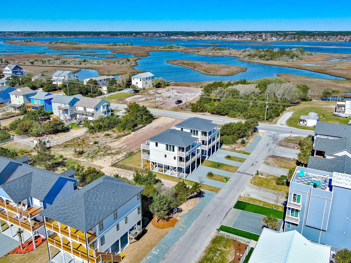 Aerial view of a coastal neighborhood featuring elevated beach homes, a winding marshland with blue water in the background, and a clear blue sky above.