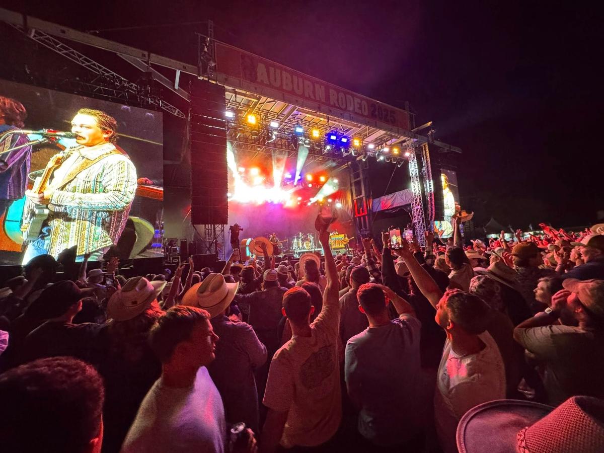 Crowd enjoying a live concert at Auburn Rodeo 2025, with bright stage lights and a band performing on a large outdoor stage. Fans raise their hands and phones, capturing the energy of the night music event. The photo shows the lively atmosphere of a popular rodeo concert experience.