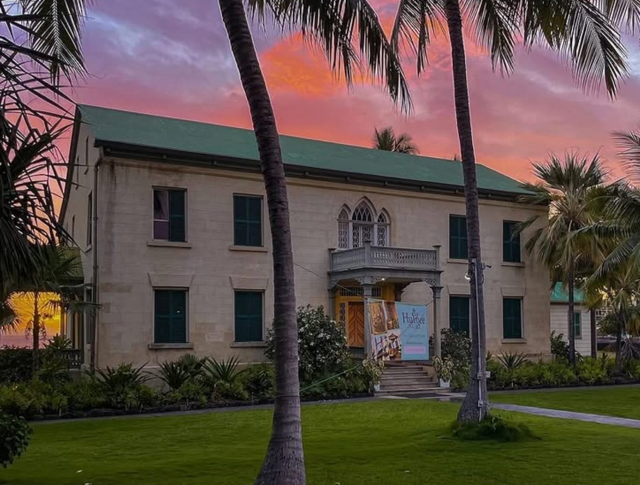 A historic stone building with green shutters and a small balcony stands framed by palm trees, set against a vibrant sunset sky in shades of pink and orange.
