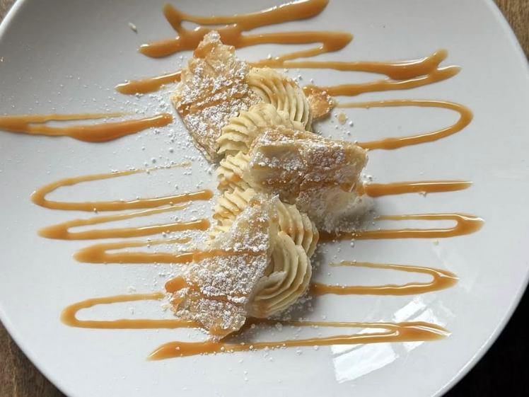 A dessert of piped cream and flaky pastry dusted with powdered sugar is artfully plated with caramel drizzle on a white dish.