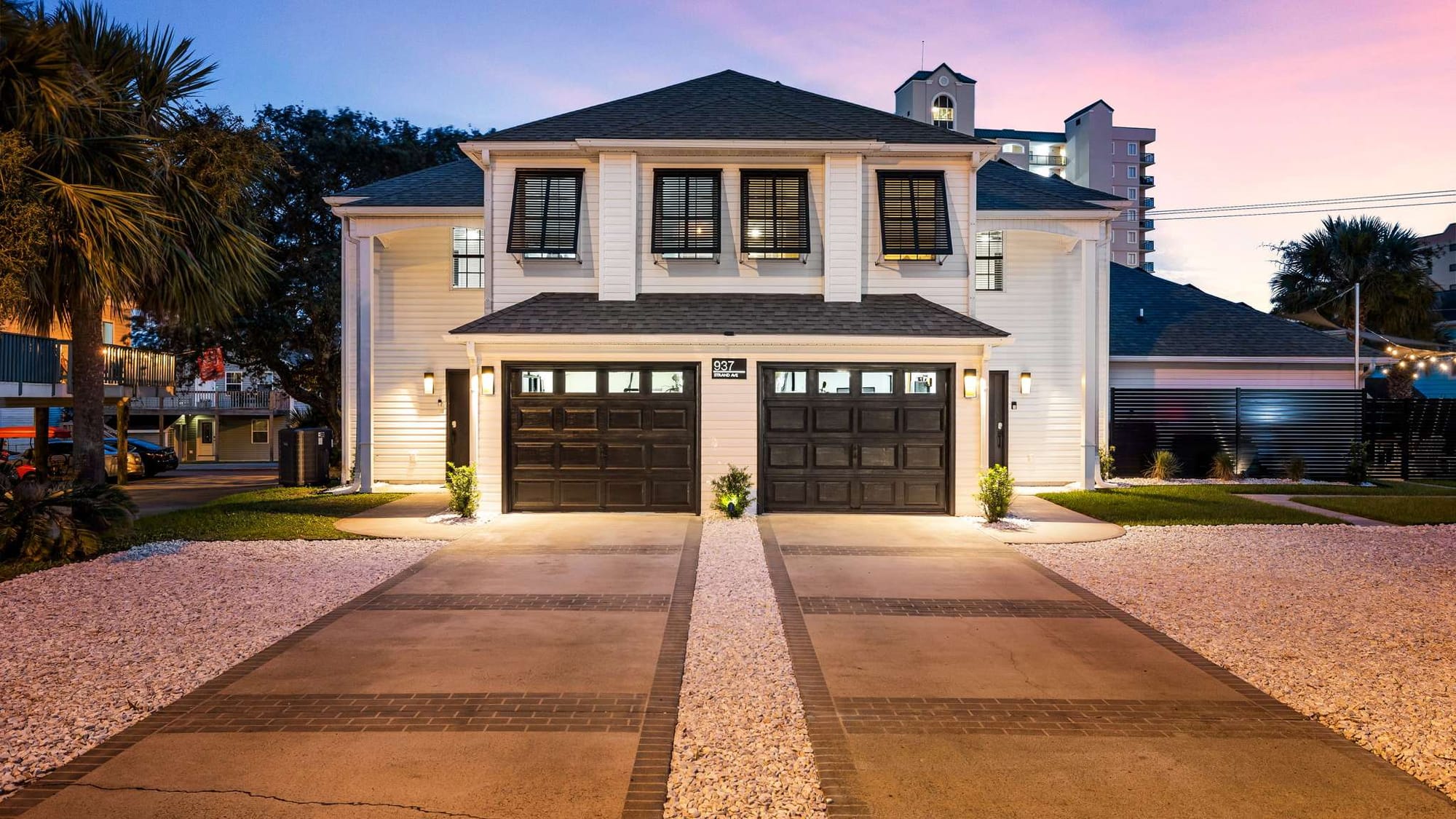 A modern two-story home with white siding, black shutters, and twin garage doors, warmly lit at dusk with a pink and blue sunset sky.