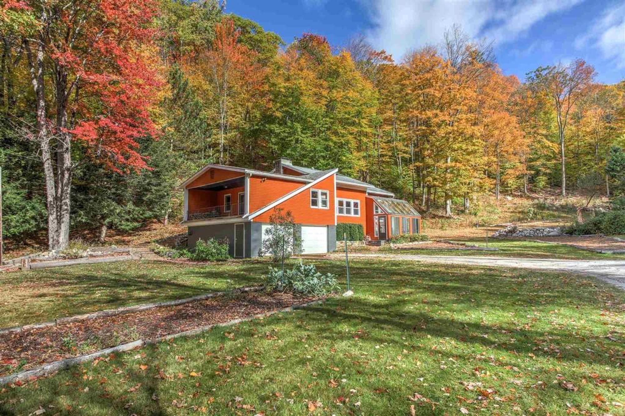 A bold orange and gray house with a sunroom entryway and a covered upper deck, set against a vibrant autumn forest with colorful red, orange, and yellow foliage.