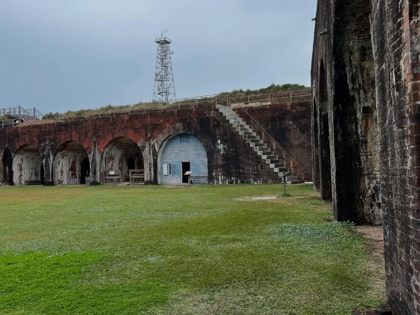 An old brick fort with weathered archways, grassy courtyard, and exterior stairs stands beneath a cloudy sky, hinting at its historic military past.