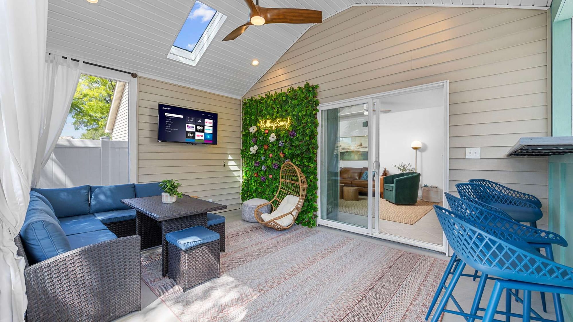 A stylish covered patio with a wicker sectional, hanging chair, neon sign on a green plant wall, wall-mounted TV, and bar seating under a skylight.