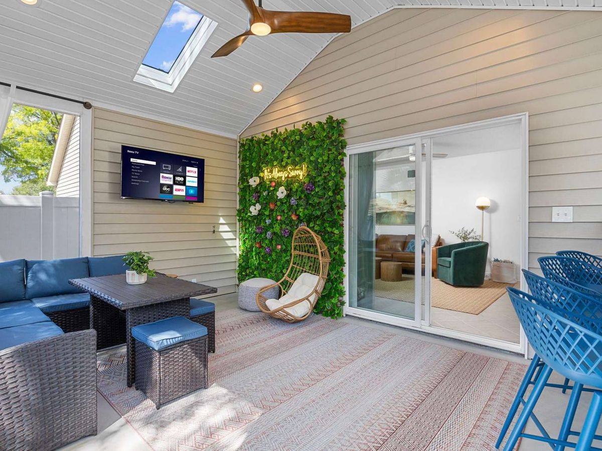 A stylish covered patio with a wicker sectional, hanging chair, neon sign on a green plant wall, wall-mounted TV, and bar seating under a skylight.
