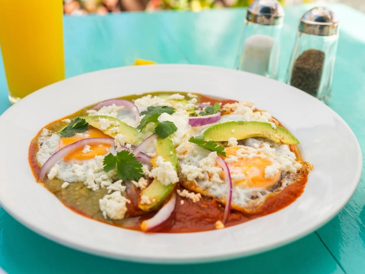 A vibrant plate of huevos rancheros topped with fried eggs, avocado slices, red onion, cilantro, crumbled cheese, and red and green salsas, served on a turquoise table with a glass of orange juice in the background.