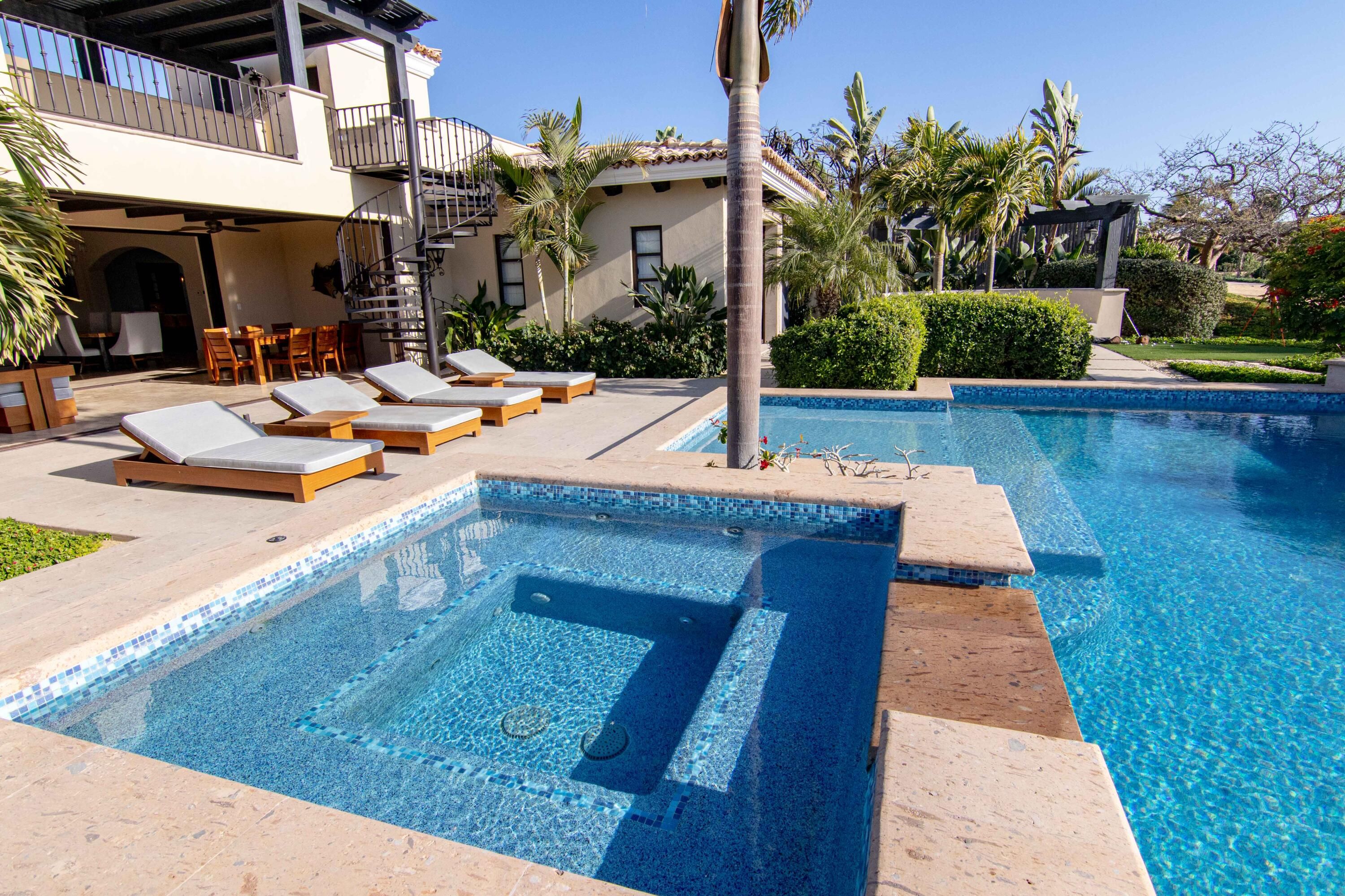 Luxury villa patio with cushioned loungers, a private hot tub and pool, tropical landscaping, and an outdoor spiral staircase leading to an upper terrace.