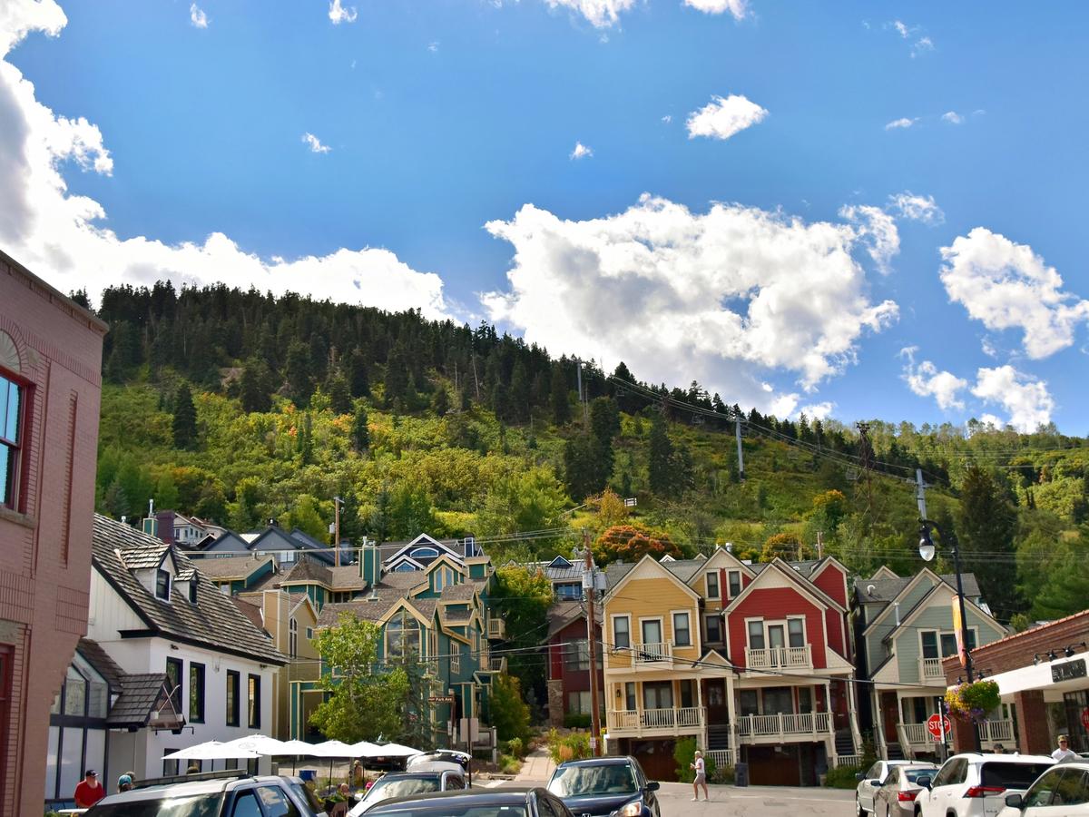 Colorful houses line a sunny street at the base of a green, tree-covered mountain with a ski lift visible under a bright blue sky dotted with fluffy white clouds.