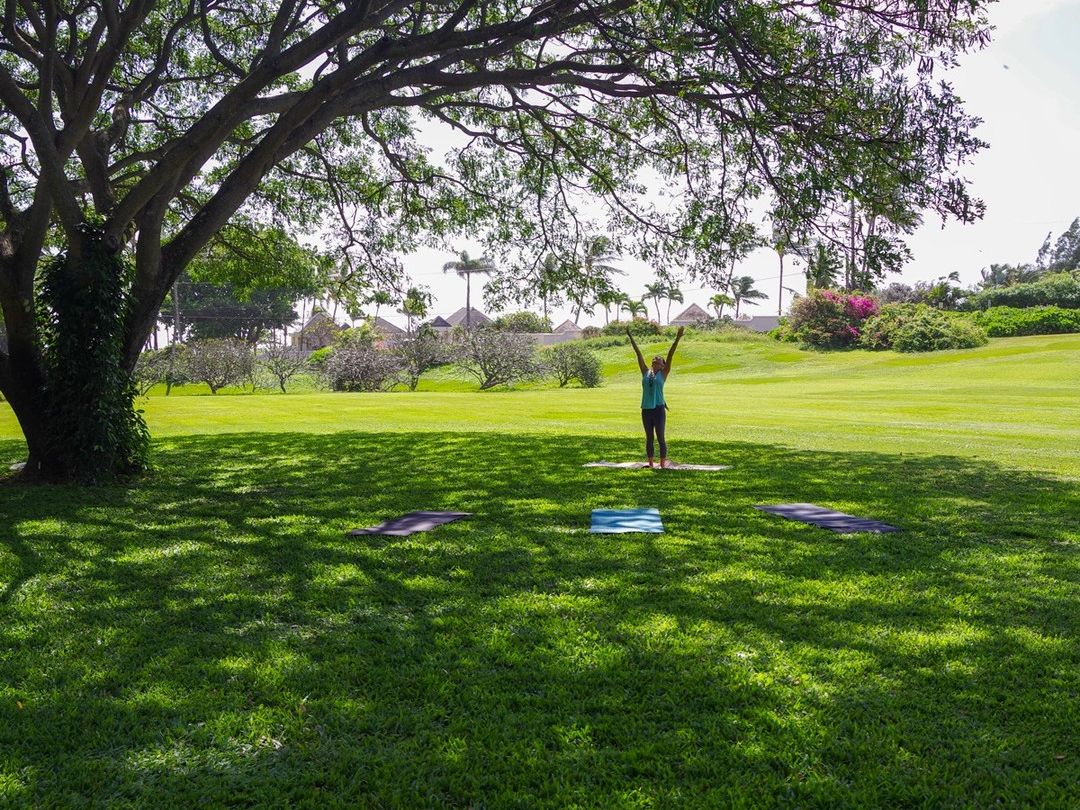 A woman practices yoga on a mat beneath a sprawling tropical tree on the lush green lawns of McBryde Allerton Garden in Kauai, Hawaii.