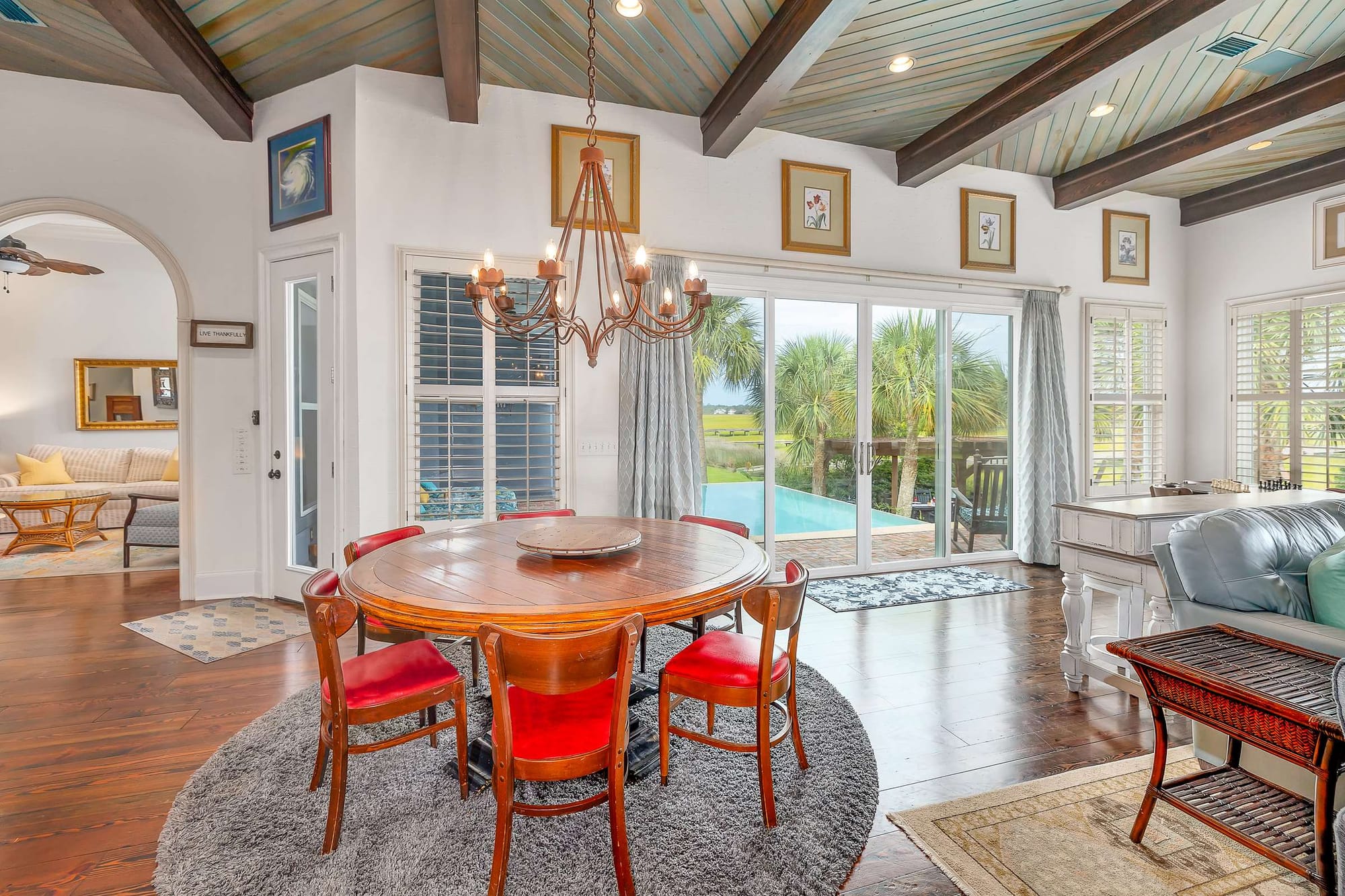 An open-concept dining and living area showcases vaulted wood beam ceilings, a wrought-iron chandelier, and a round wooden table with red leather chairs, leading out through sliding glass doors to a palm-lined infinity pool and marsh view. The warm coastal decor and abundant natural light create an inviting gathering space for large groups.