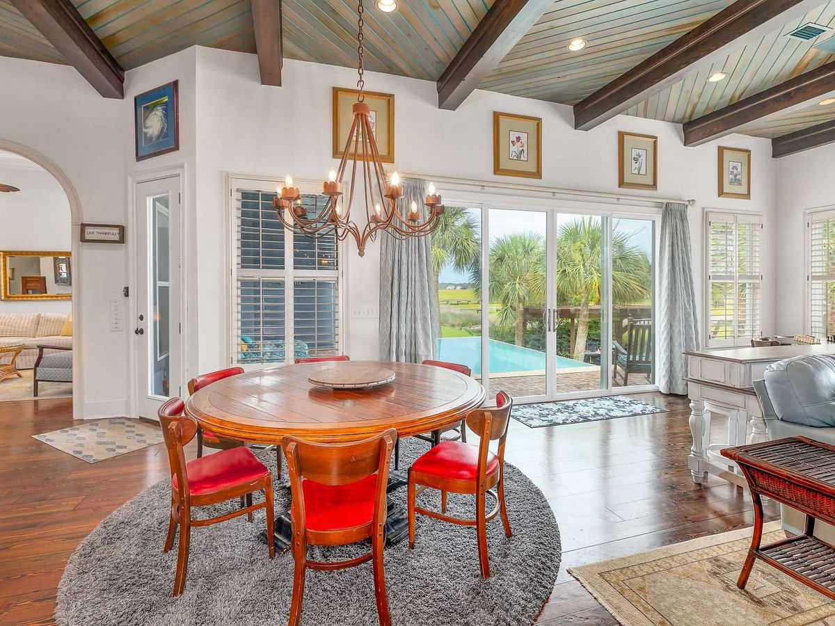 An open-concept dining and living area showcases vaulted wood beam ceilings, a wrought-iron chandelier, and a round wooden table with red leather chairs, leading out through sliding glass doors to a palm-lined infinity pool and marsh view. The warm coastal decor and abundant natural light create an inviting gathering space for large groups.