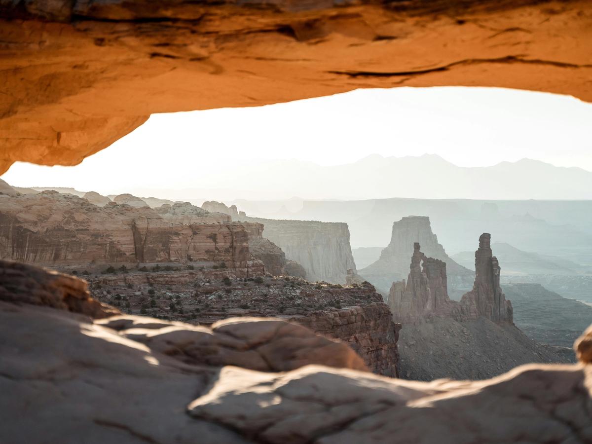 A stunning desert landscape is viewed through a natural sandstone arch, revealing dramatic rock spires and mesas bathed in soft sunlight.