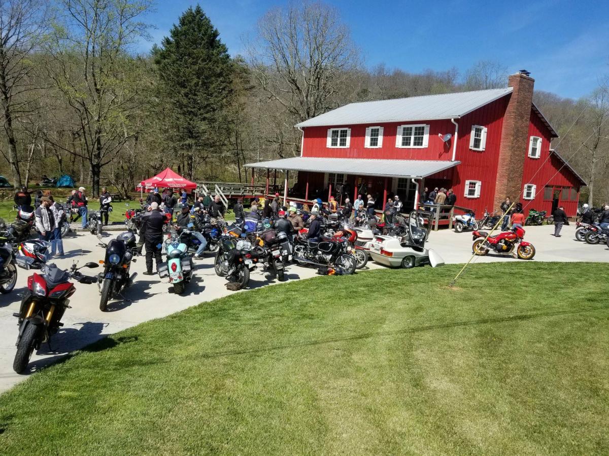 A group of motorcycles is parked outside a bright red barn where riders gather for food and camaraderie. The sunny day and open countryside create the perfect pit stop for road adventurers.