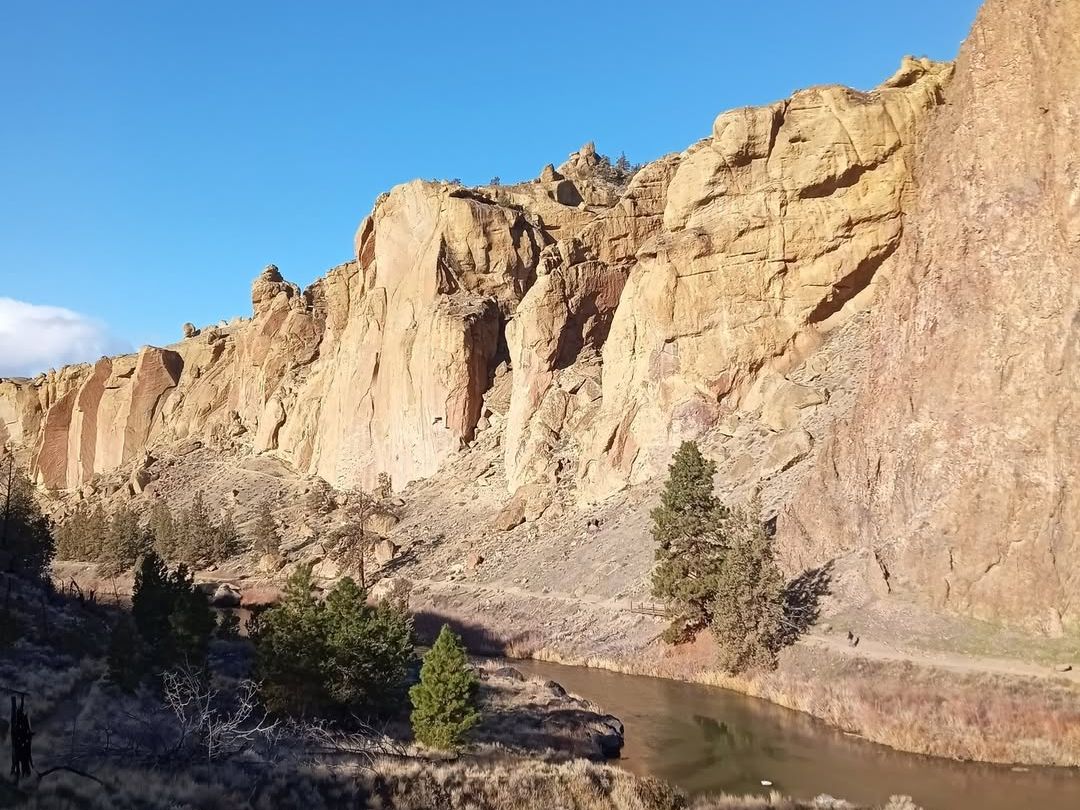 Towering golden cliffs rise above a winding river, set against a backdrop of clear blue skies and high desert scenery. A stunning view highlighting the rugged beauty near the Sunriver Nature Center.