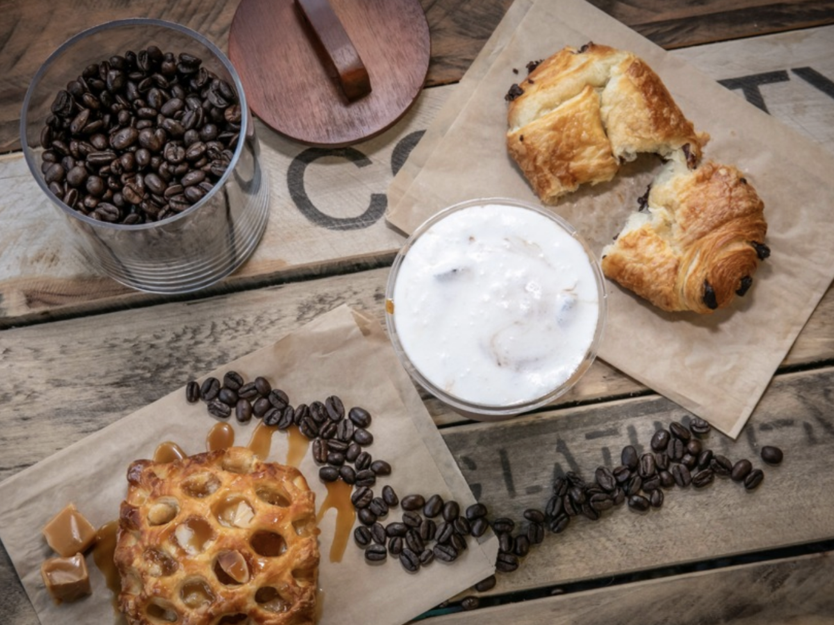 Overhead view of a rustic wooden table with a chocolate croissant, a caramel pastry with cubes of caramel and scattered coffee beans, a creamy iced coffee, and a jar filled with whole coffee beans.