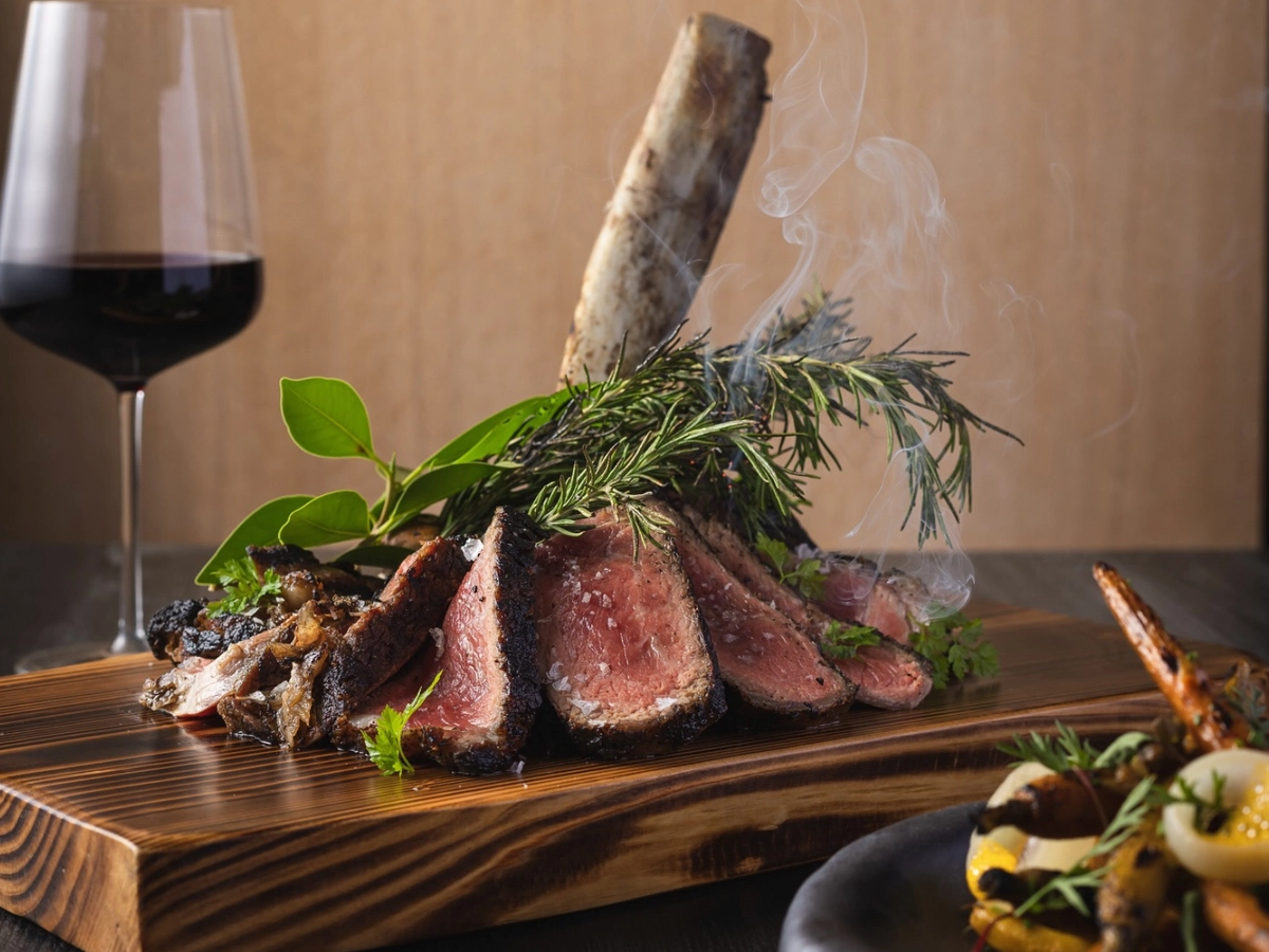 Slices of perfectly seared rare steak are served on a wooden board with herbs and roasted mushrooms, accompanied by a glass of red wine and a side of colorful roasted vegetables.