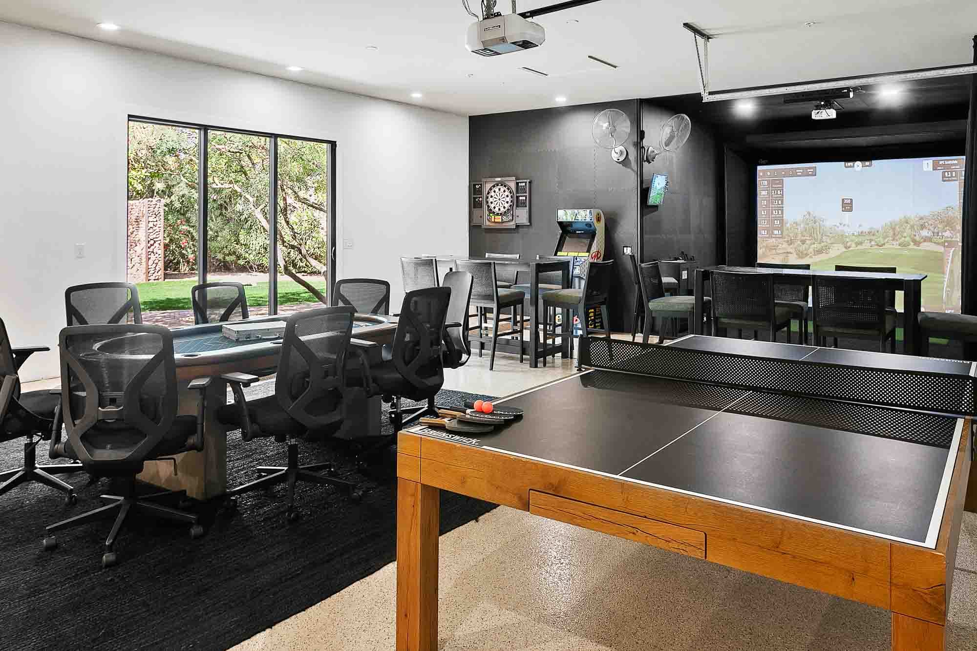 Modern entertainment room featuring a ping pong table, poker table with office-style chairs, high-top seating, arcade game, dartboard, and a golf simulator area with large screen.