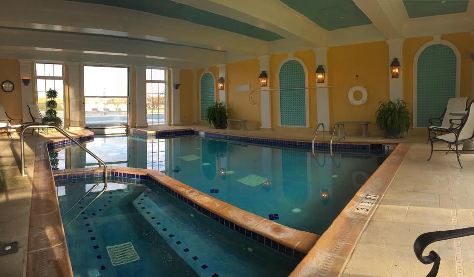 An elegant indoor pool at Keswick Hall features blue-tiled lanes beneath a coffered ceiling painted in yellow and teal. Arched alcoves line the walls, flanked by wrought-iron lanterns and potted palms, with large windows overlooking the grounds.