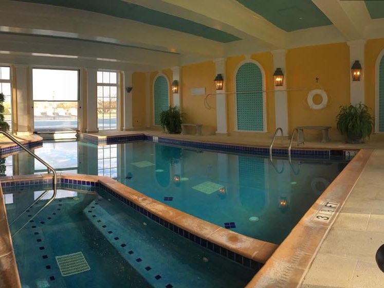 An elegant indoor pool at Keswick Hall features blue-tiled lanes beneath a coffered ceiling painted in yellow and teal. Arched alcoves line the walls, flanked by wrought-iron lanterns and potted palms, with large windows overlooking the grounds.
