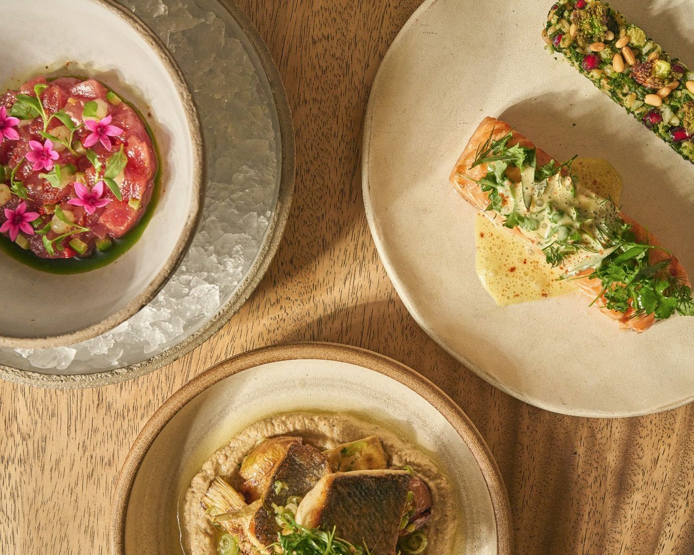 A refined set of Mediterranean dishes sits on a wooden table, featuring fresh seafood, herbs, and bright garnishes. The plates look elegant and thoughtfully crafted, with pops of color from edible flowers and greens. It captures a calm, upscale dining experience.