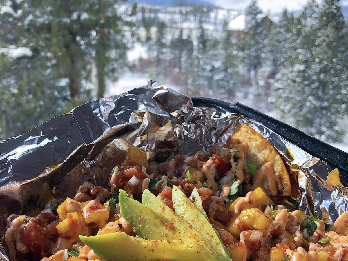 Close-up of a taco filled with diced seafood, salsa, and avocado slices, served in foil with a snowy forest and mountain landscape in the background.