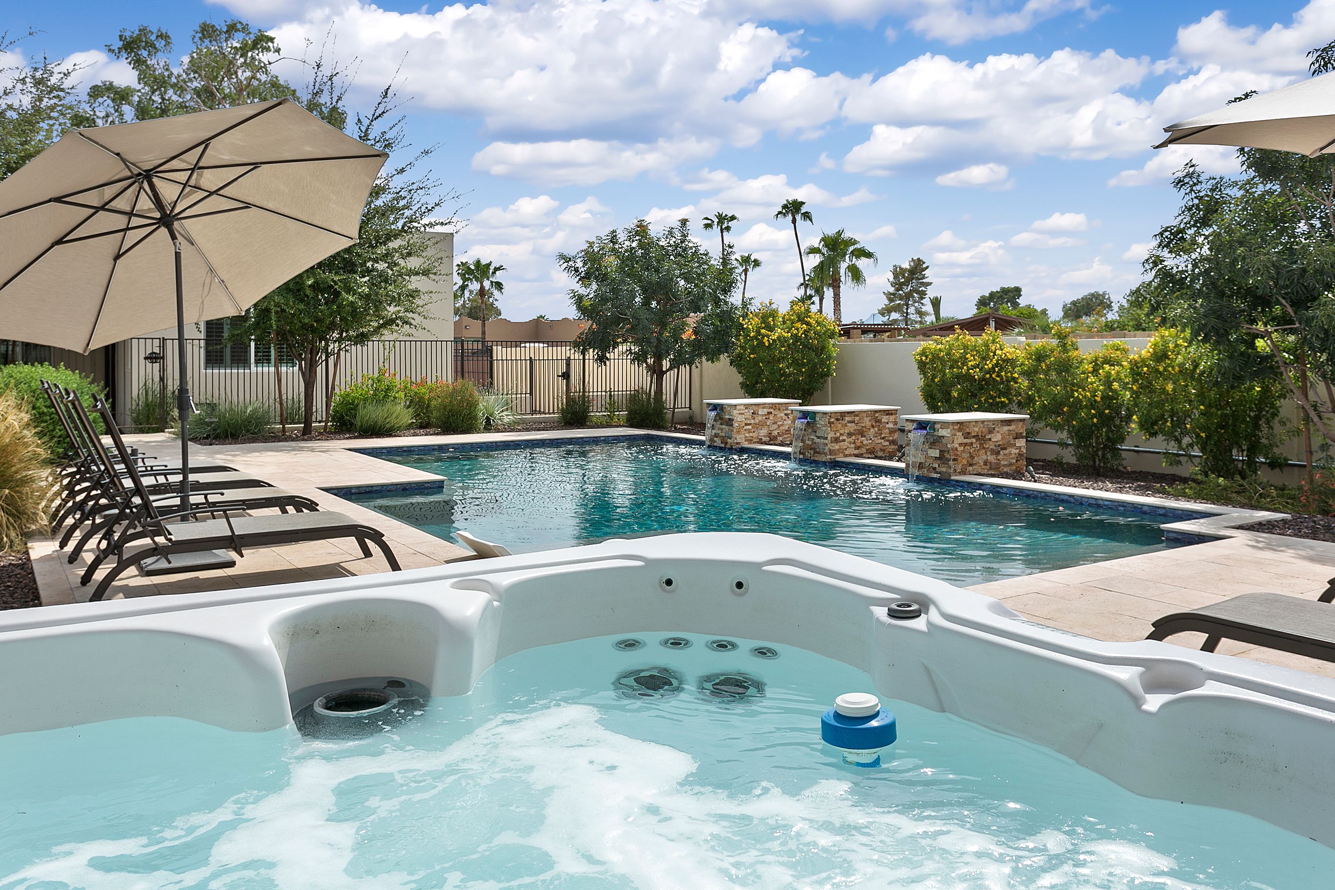 A private resort-style backyard featuring a sparkling pool with cascading water features, an inviting hot tub, and shaded lounge seating surrounded by lush greenery.