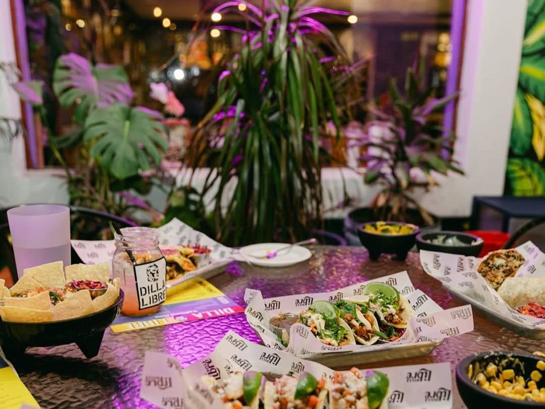 A lively table at Dilla Libre Dos with tacos, chips, and sides, set against a neon sign and tropical décor. The fun and vibrant vibe makes this spot a go-to for creative Mexican eats.