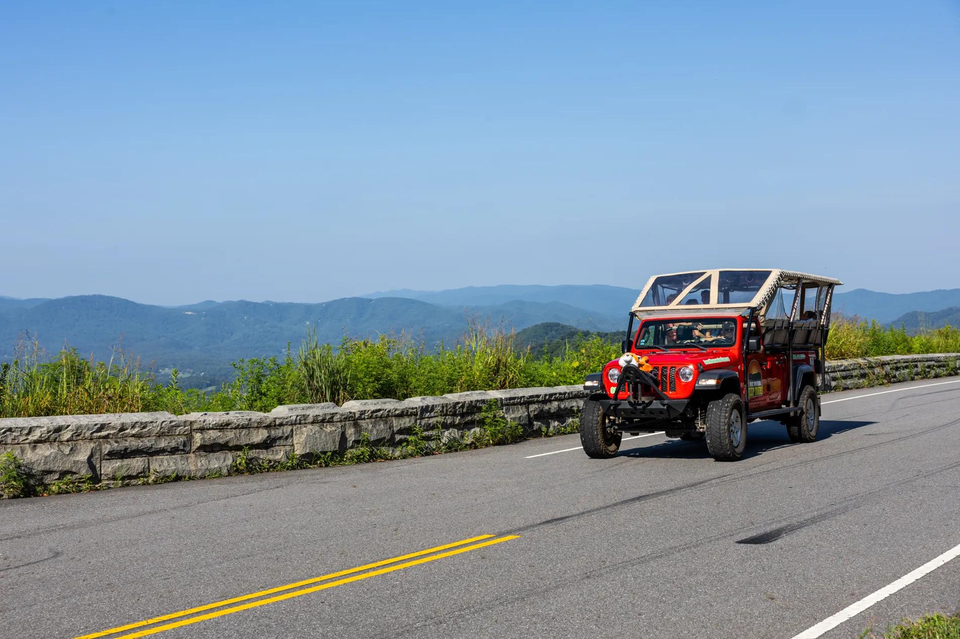 : A red open-top Jeep drives along a mountain road overlooking rolling blue peaks of the Smoky Mountains. Smoky Mountain Jeep Tours offers guided off-road adventures through stunning scenic routes.