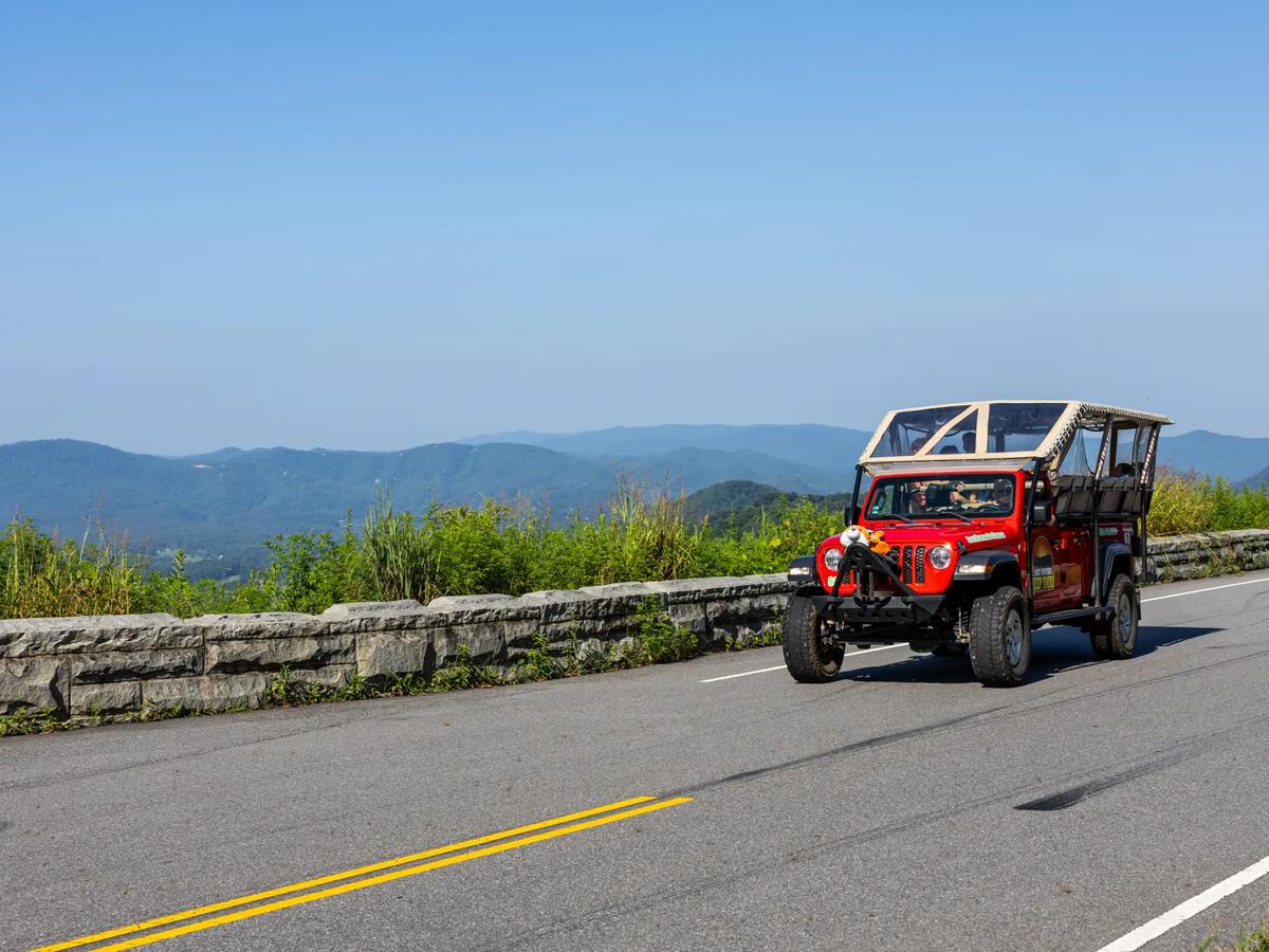 : A red open-top Jeep drives along a mountain road overlooking rolling blue peaks of the Smoky Mountains. Smoky Mountain Jeep Tours offers guided off-road adventures through stunning scenic routes.