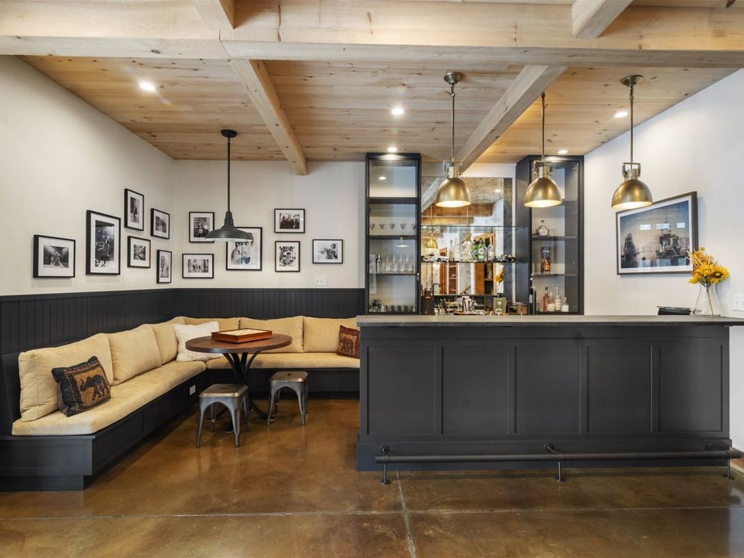 A stylish basement bar area at Owls Head Estate features a dark slate bar with brass pendant lighting, a built-in liquor display, and a cozy L-shaped banquette with a gallery wall of black and white photographs above wainscoting. The exposed natural wood beam ceiling adds warmth to the sophisticated entertainment space.