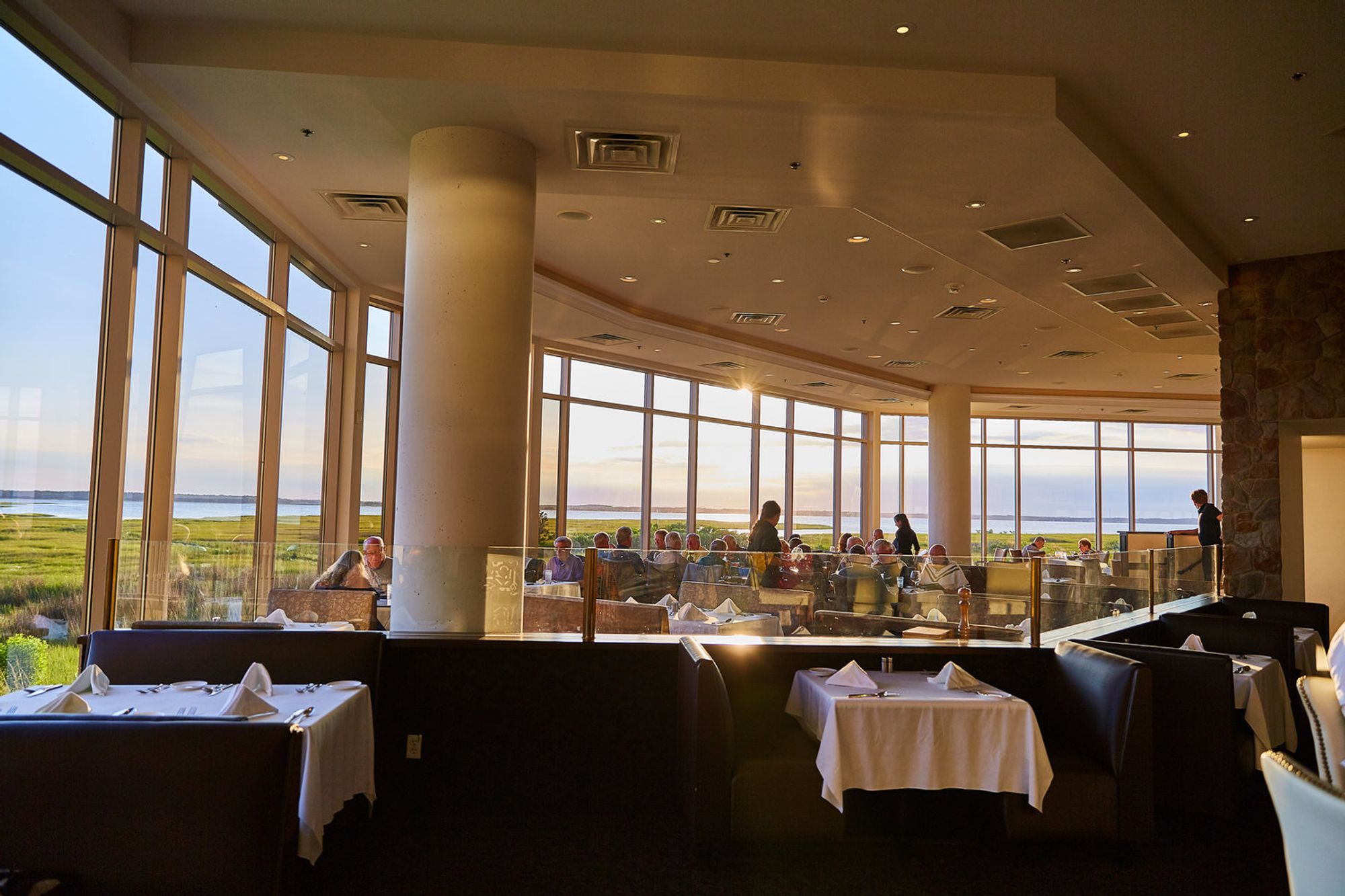 Golden hour sunlight pouring into The Hobbit Restaurant, with coastal marsh views and guests dining by the window. A serene and scenic evening setting in Ocean City.