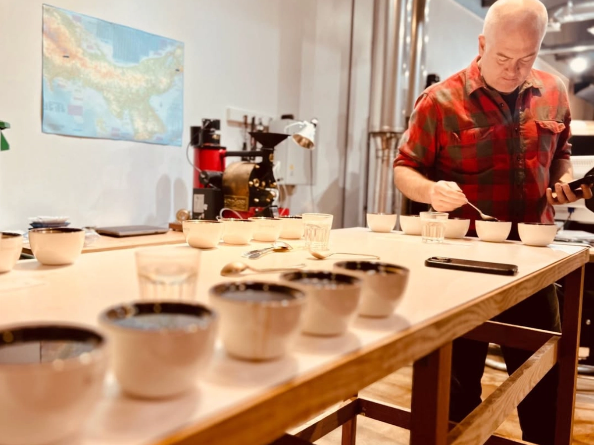 A man in a red plaid shirt participates in a coffee cupping session at a specialty coffee roastery. He uses a spoon to taste from a row of cupping bowls on a wooden table. The background features coffee roasting equipment, a map on the wall, and an industrial-style setting with exposed pipes and lighting.