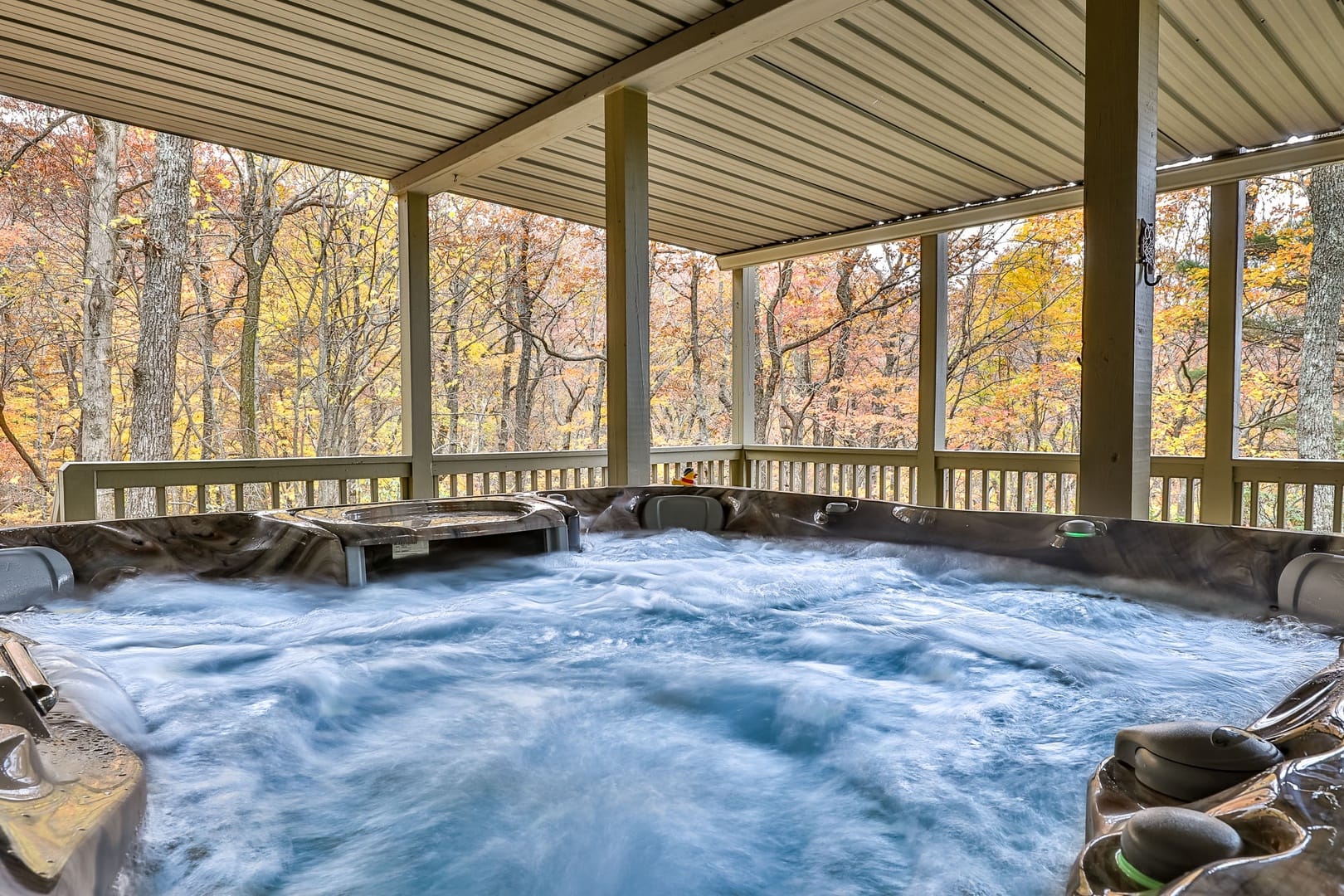 Covered outdoor hot tub with bubbling water on a screened cabin deck, surrounded by autumn forest views with golden and rust-colored leaves, offering a cozy, private mountain retreat setting.