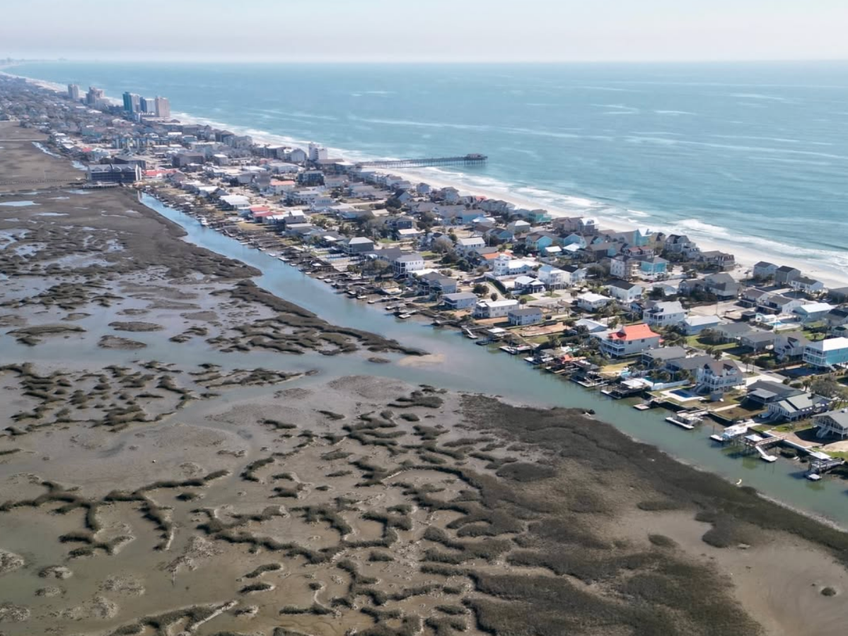 A sweeping aerial view of a Myrtle Beach barrier island, with oceanfront homes and sandy beaches on one side and winding tidal marshes and waterways on the other, stretching toward the pier and skyline in the distance.