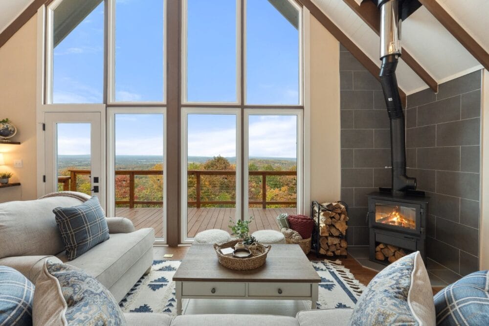 Cozy living room with vaulted ceilings and floor-to-ceiling windows overlooking a mountain view, featuring a wood-burning stove, stacked firewood, and plush sofas with blue plaid pillows.