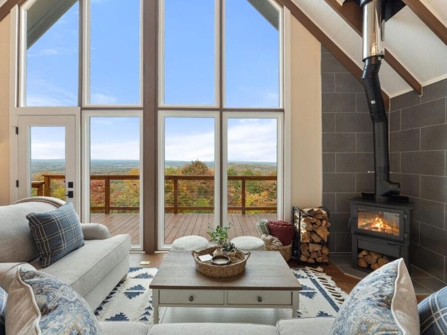 Cozy living room with vaulted ceilings and floor-to-ceiling windows overlooking a mountain view, featuring a wood-burning stove, stacked firewood, and plush sofas with blue plaid pillows.