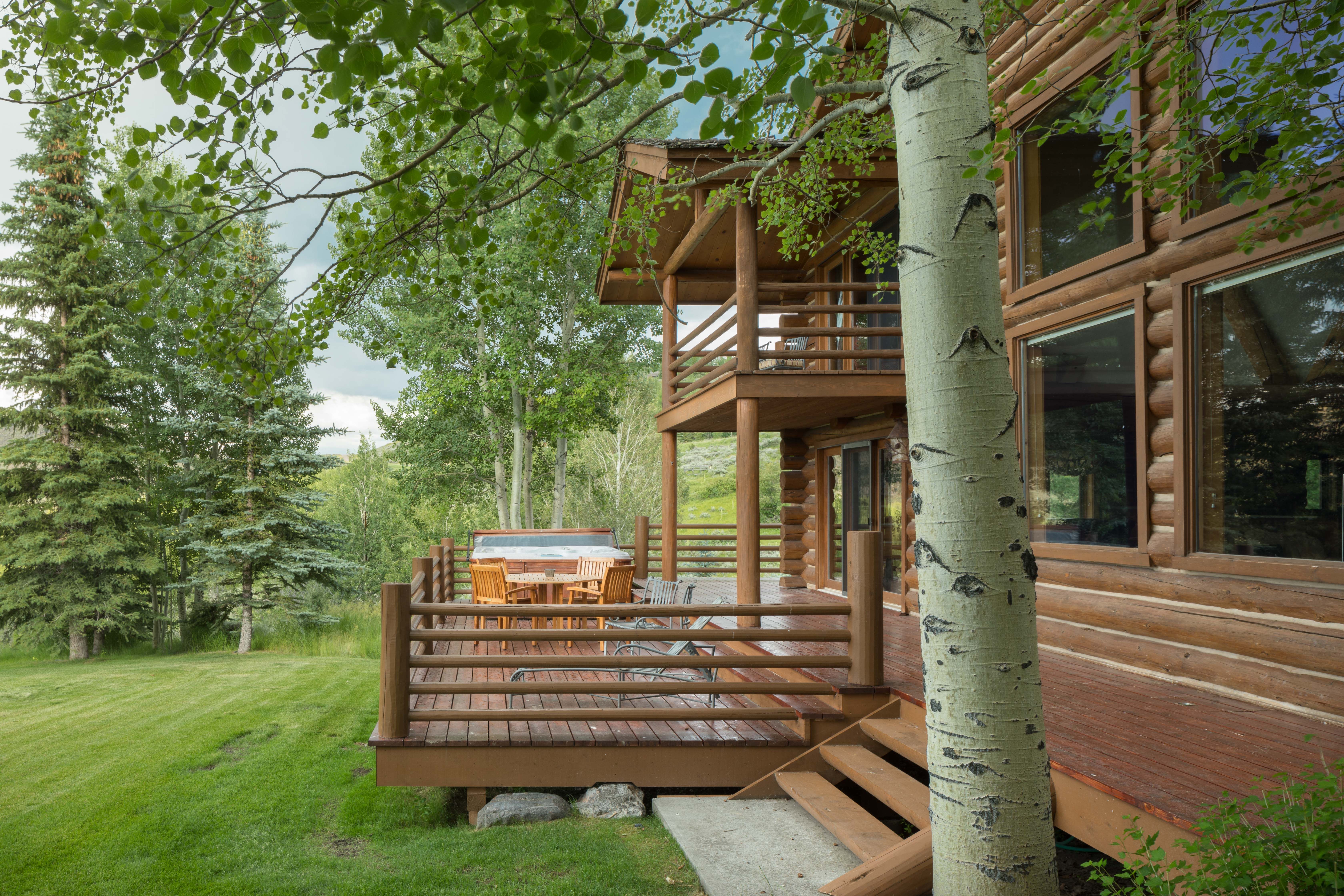 Wooden deck of a log cabin nestled in a lush forest, featuring outdoor dining furniture and a hot tub overlooking a grassy yard surrounded by tall pine and aspen trees.