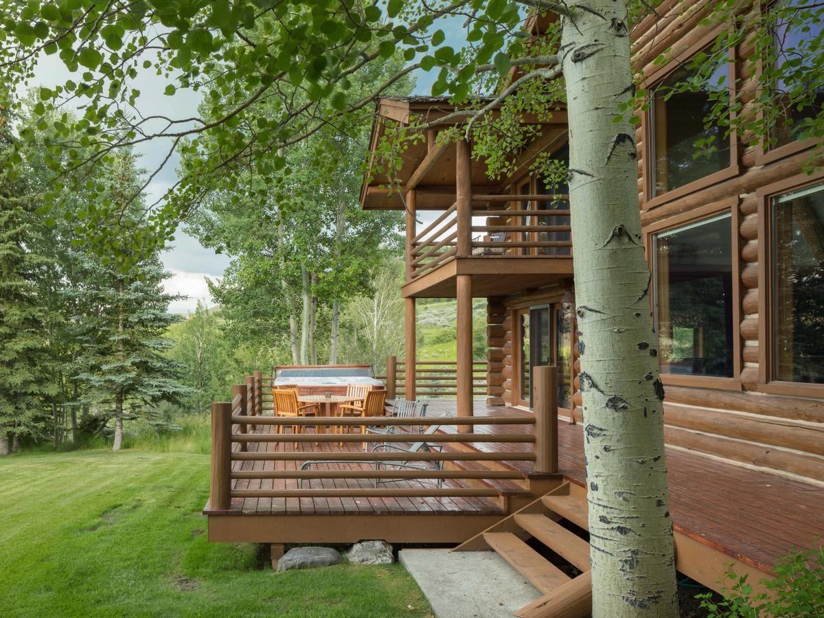 Wooden deck of a log cabin nestled in a lush forest, featuring outdoor dining furniture and a hot tub overlooking a grassy yard surrounded by tall pine and aspen trees.