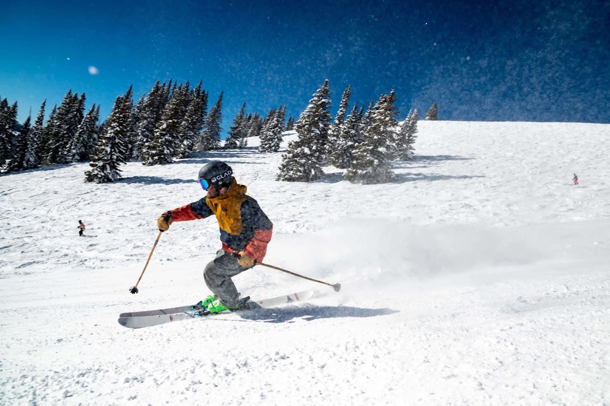 A skier carves down a fresh powder slope under bright blue skies at a winter resort. Snow sprays into the air, capturing the excitement and energy of a day on the mountain.