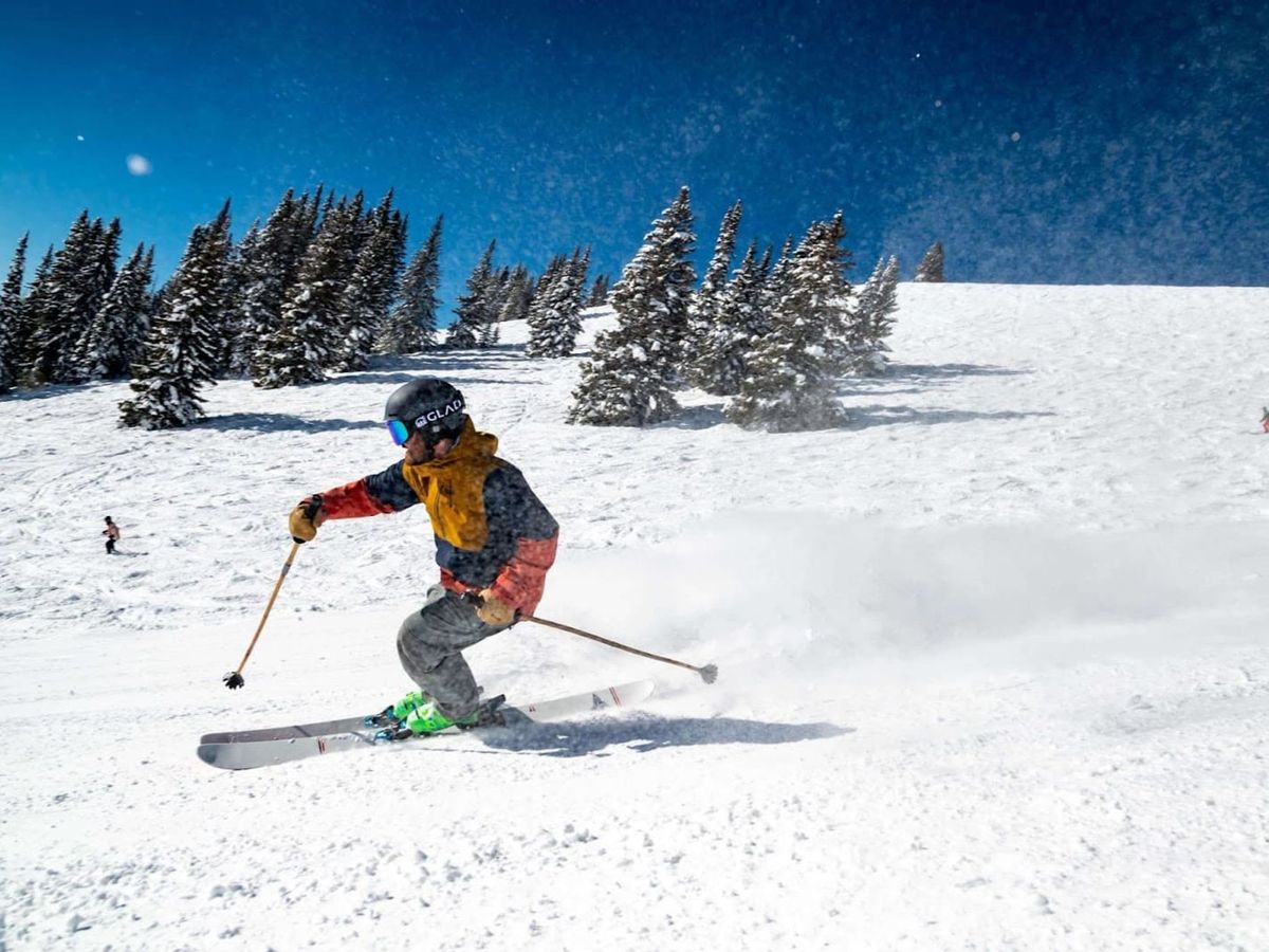 A skier carves down a fresh powder slope under bright blue skies at a winter resort. Snow sprays into the air, capturing the excitement and energy of a day on the mountain.