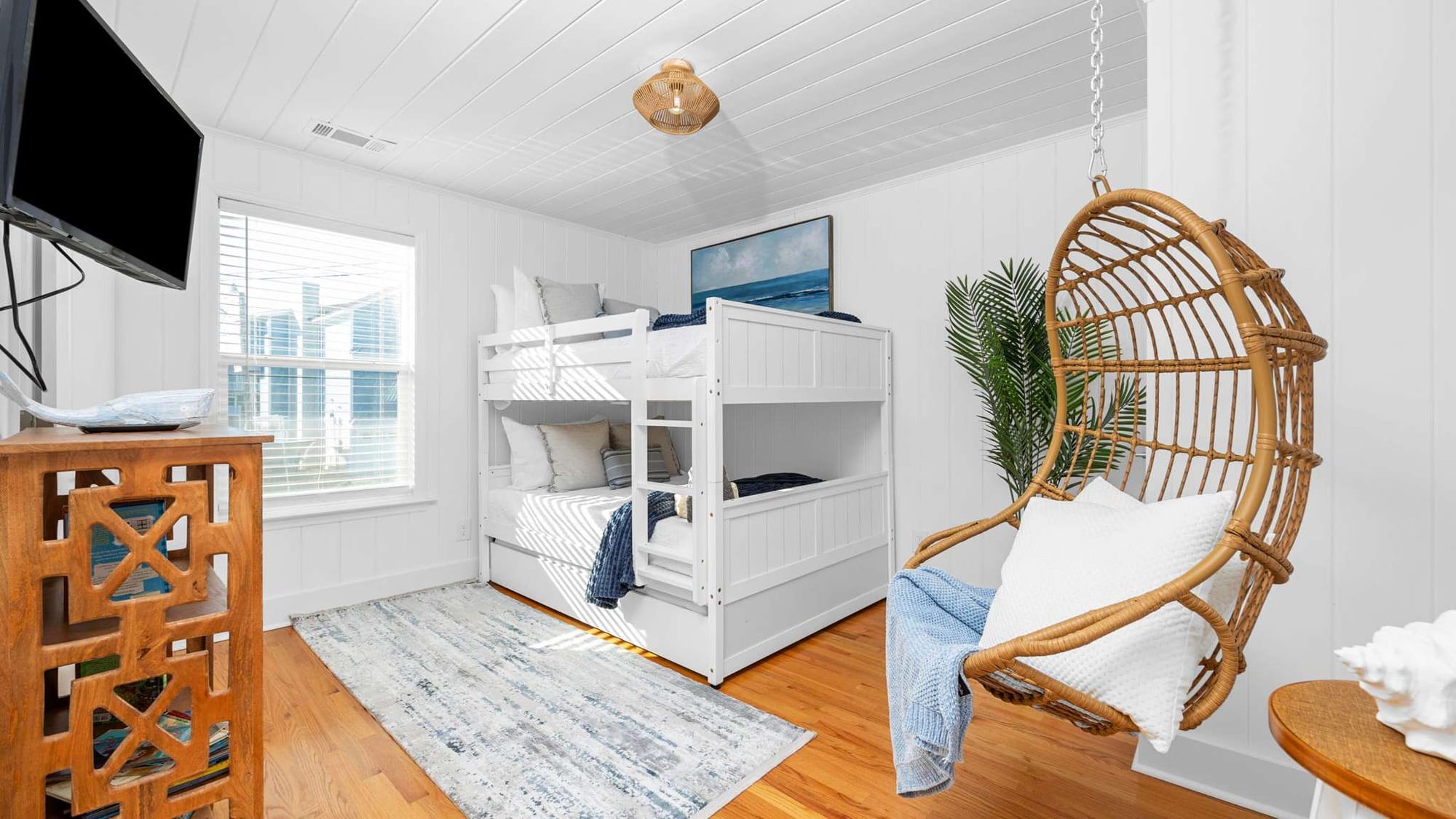 A bright coastal-style bedroom with white bunk beds, a hanging wicker chair, and natural wood accents.