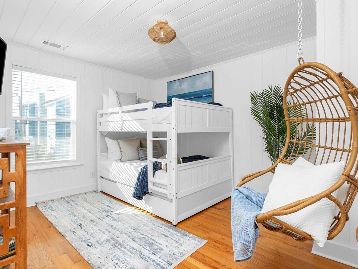 A bright coastal-style bedroom with white bunk beds, a hanging wicker chair, and natural wood accents.