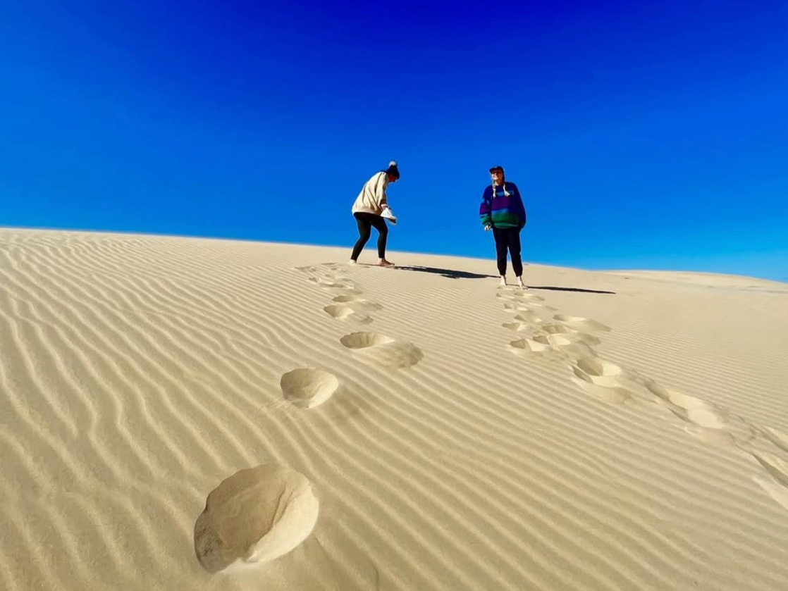 two people side by side on mound of sand with footsteps in the sand